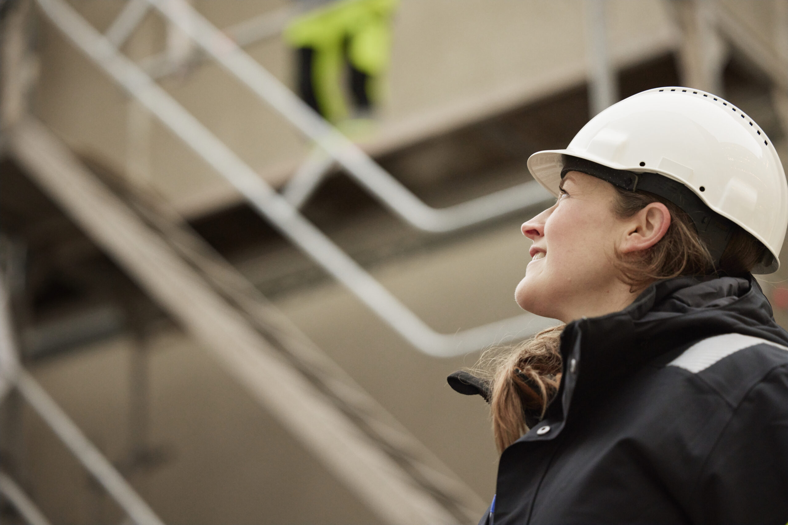Kvinna i skyddshjälm i industrimiljö blickar upp – symboliserar det operativa arbetet med att identifiera utrustning som kan återbrukas i cirkulärt inköpsarbete.
