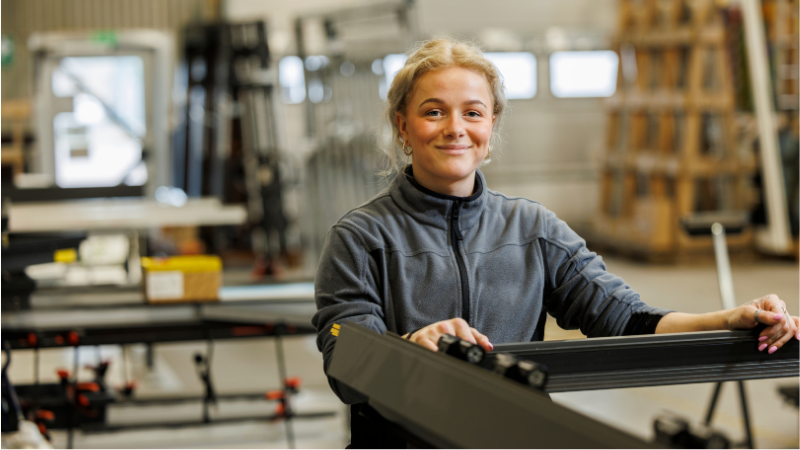 En kvinnlig industrimedarbetare i verkstadsmiljö ler mot kameran, symboliserar kontroll och resurseffektivitet i produktionen.